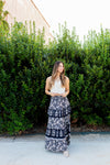 Woman wearing a white top and navy floral skirt standing in front of green bushes.