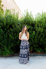Woman wearing a white top and navy floral skirt standing in front of green bushes.