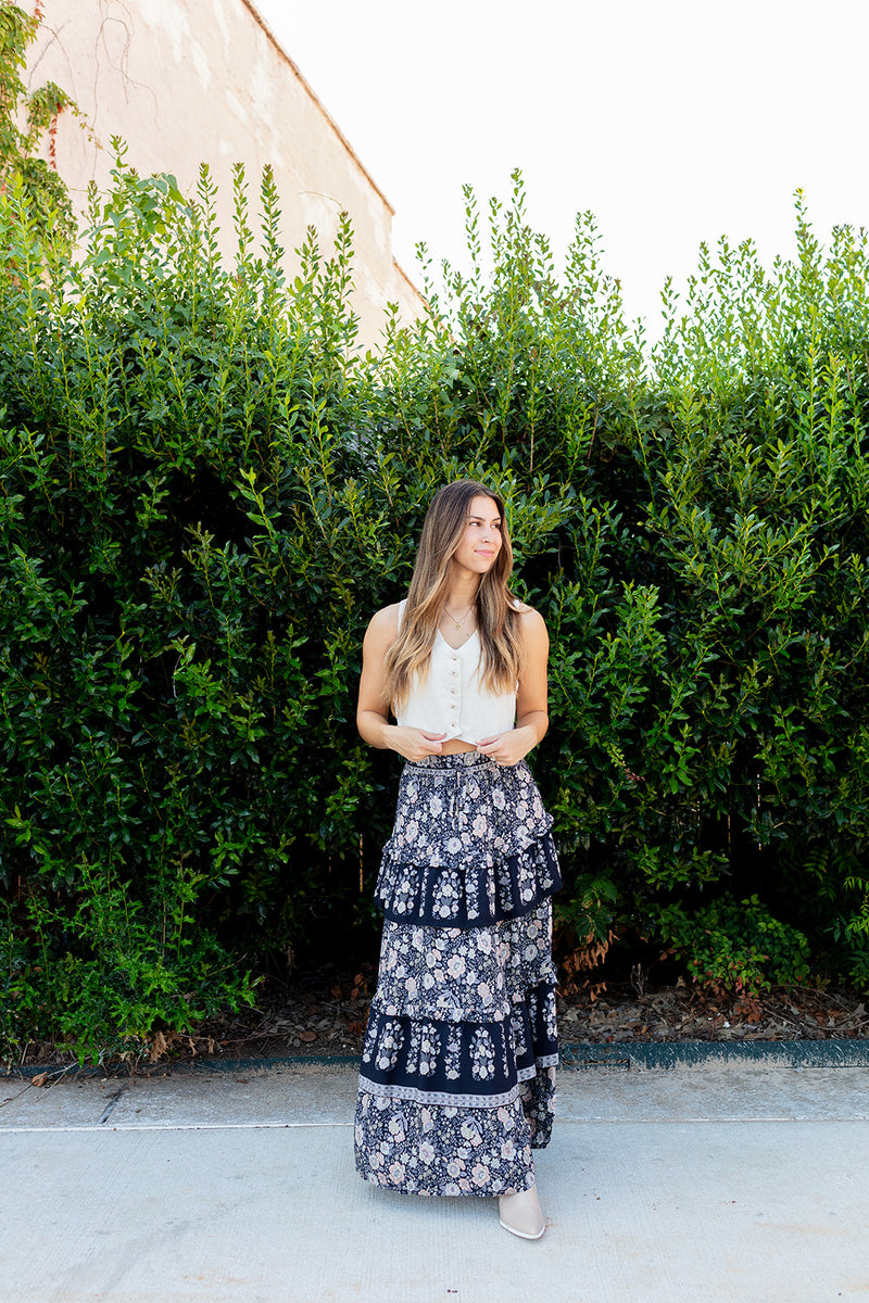 Woman wearing a white top and navy floral skirt standing in front of green bushes.
