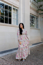 Woman wearing a long-sleeve floral maxi dress with smocked bodice and tiered skirt, standing outside a historic stone building