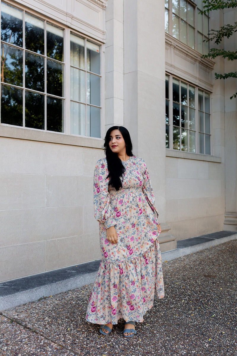 Woman wearing a long-sleeve floral maxi dress with smocked bodice and tiered skirt, standing outside a historic stone building