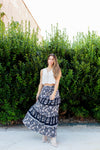 Woman in a white top and floral skirt standing in front of green bushes.