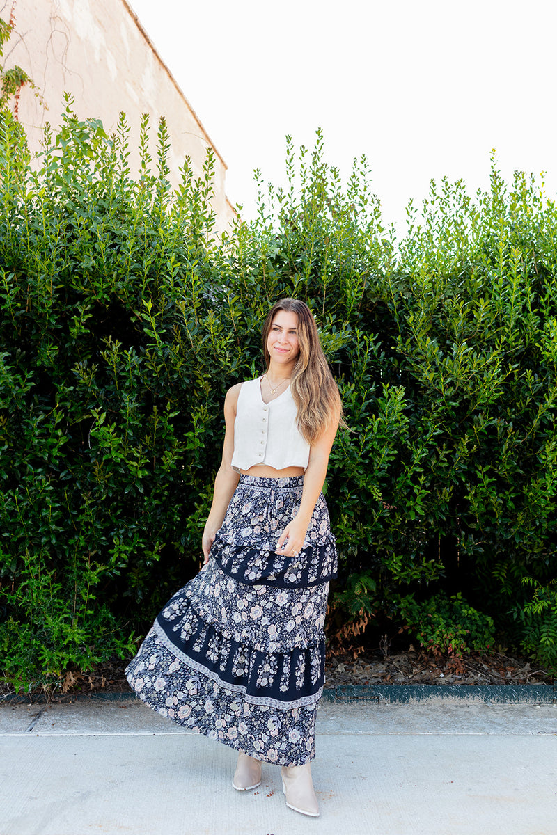 Woman in a white top and floral skirt standing in front of green bushes.