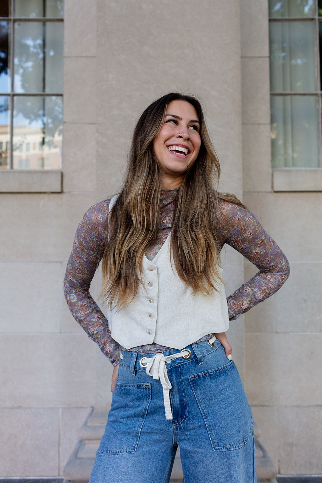 Woman wearing a sheer long-sleeve top over a white shirt and blue jeans, standing in front of a building.