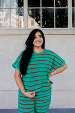 Woman wearing a green and brown striped outfit standing against a light-colored wall.
