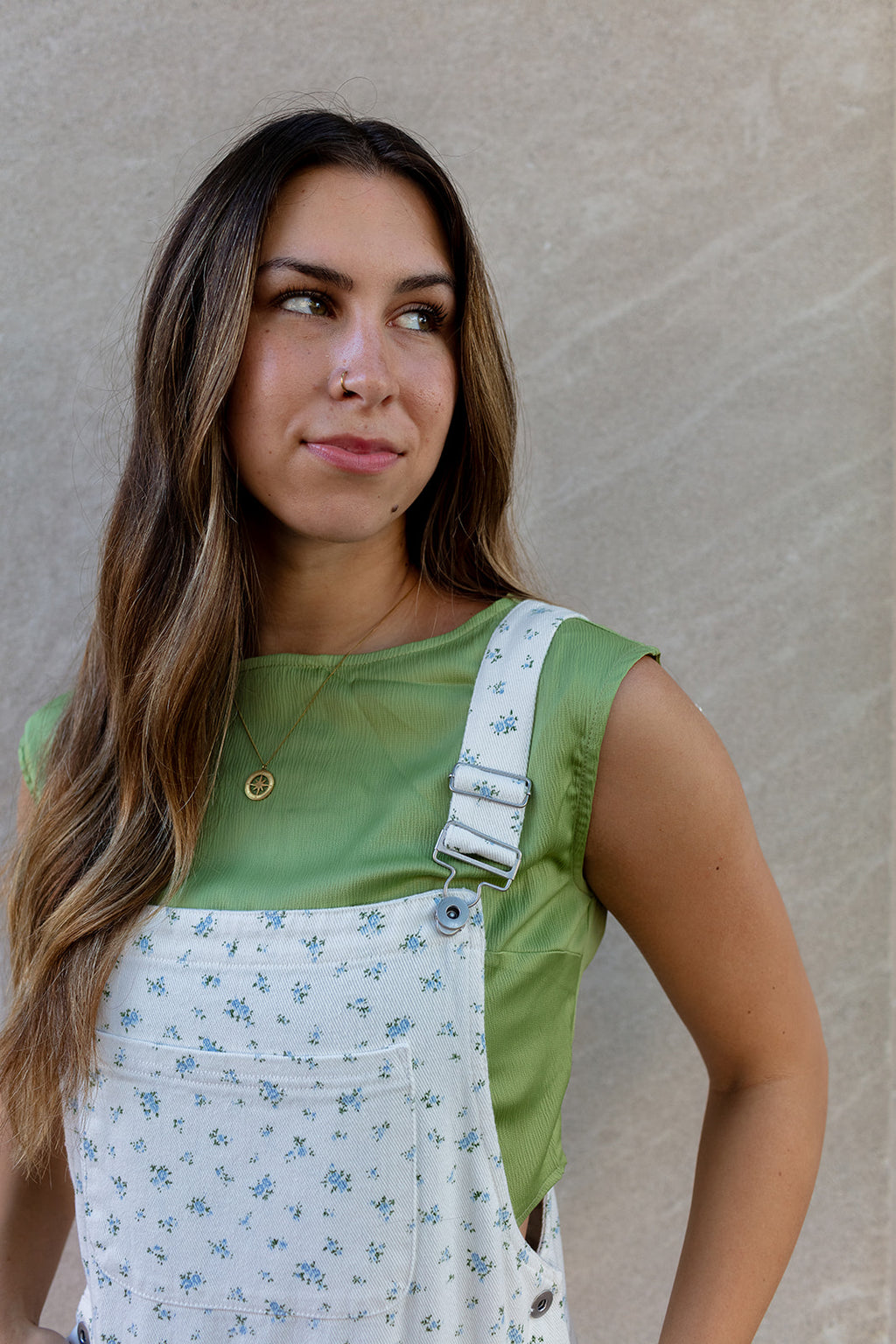 Woman wearing a green shirt and floral overalls against a beige wall