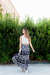 Woman wearing a white top and a long, dark floral skirt standing in front of green bushes.