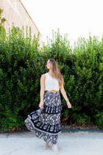 Woman wearing a white top and a long, dark floral skirt standing in front of green bushes.