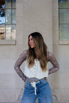 Woman wearing a floral long-sleeve top over a white shirt with blue jeans, standing against a beige building.