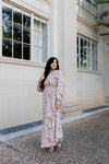 Woman in a cream floral maxi dress with long sleeves and smocked waist, posing by a stone building with large windows