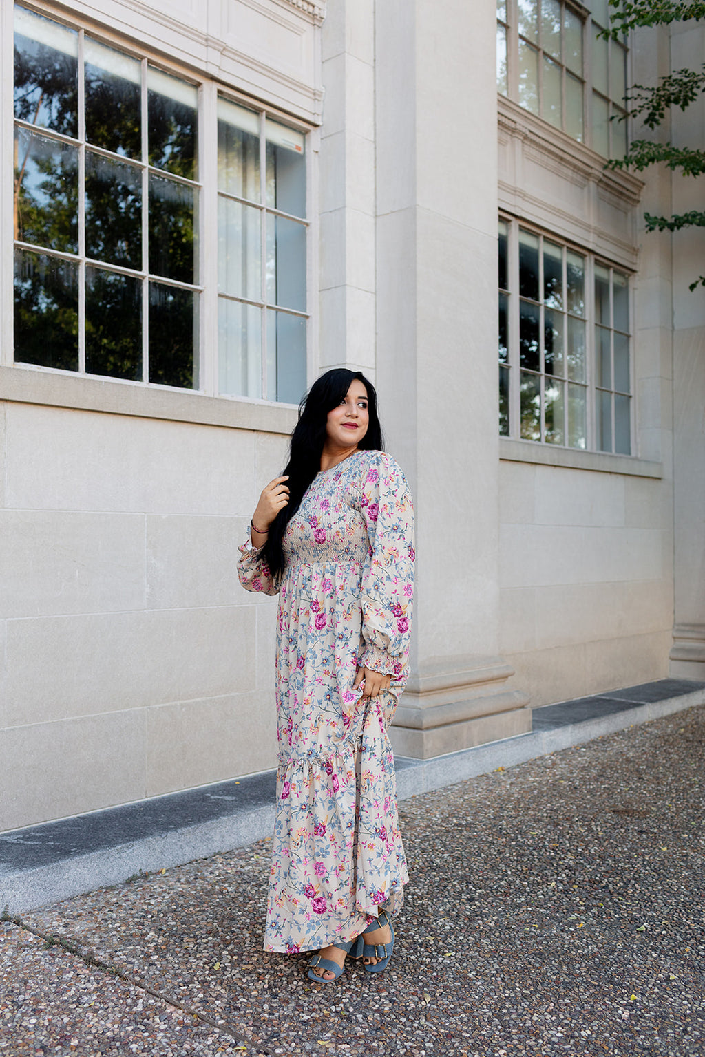 Woman in a cream floral maxi dress with long sleeves and smocked waist, posing by a stone building with large windows
