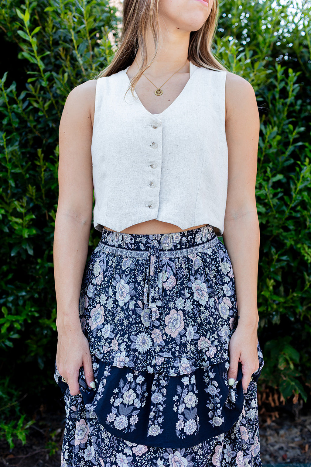 Woman wearing a white sleeveless top and floral skirt against a green bush background