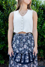 Woman wearing a white sleeveless top and floral skirt against a green bush background