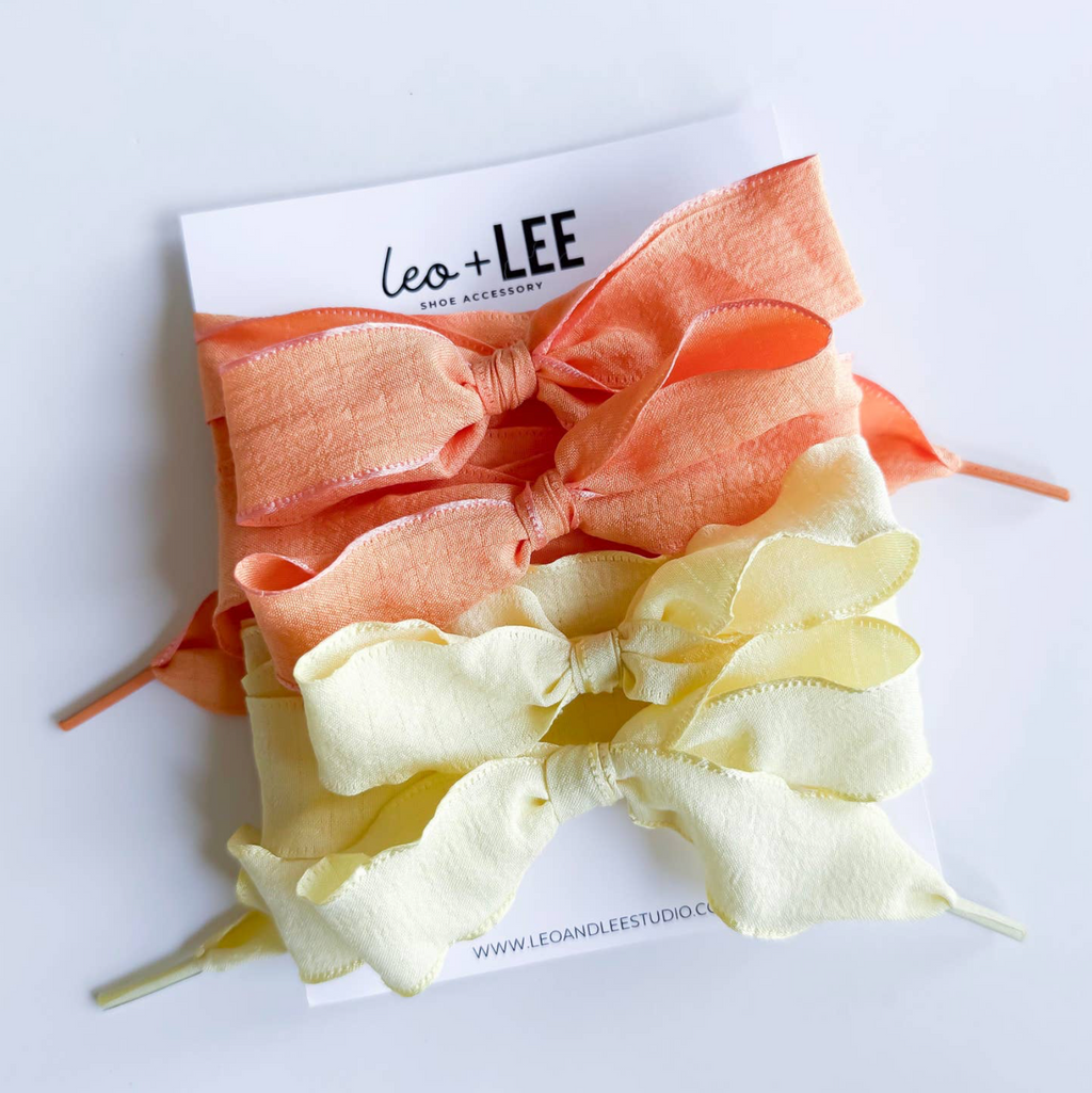 Two hair bows, one peach and one yellow, on a white background with 'Leo + Lee' branding.