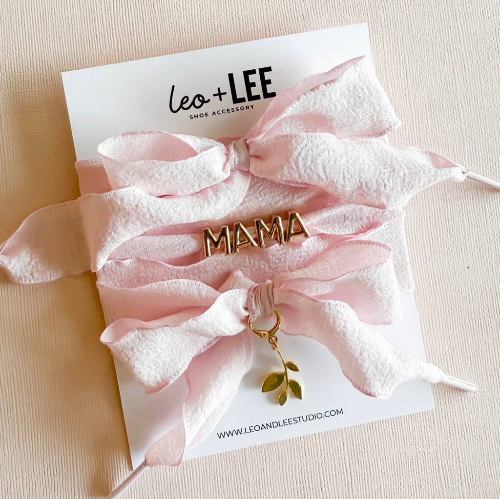 Soft blush ribbon laces featuring a gold “MAMA” charm and dainty leafy pendant 🌿, styled on luxe packaging.