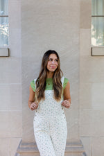Woman wearing a green top and white overalls standing against a beige wall.