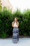 Woman in a white top and floral skirt standing in front of a green hedge.
