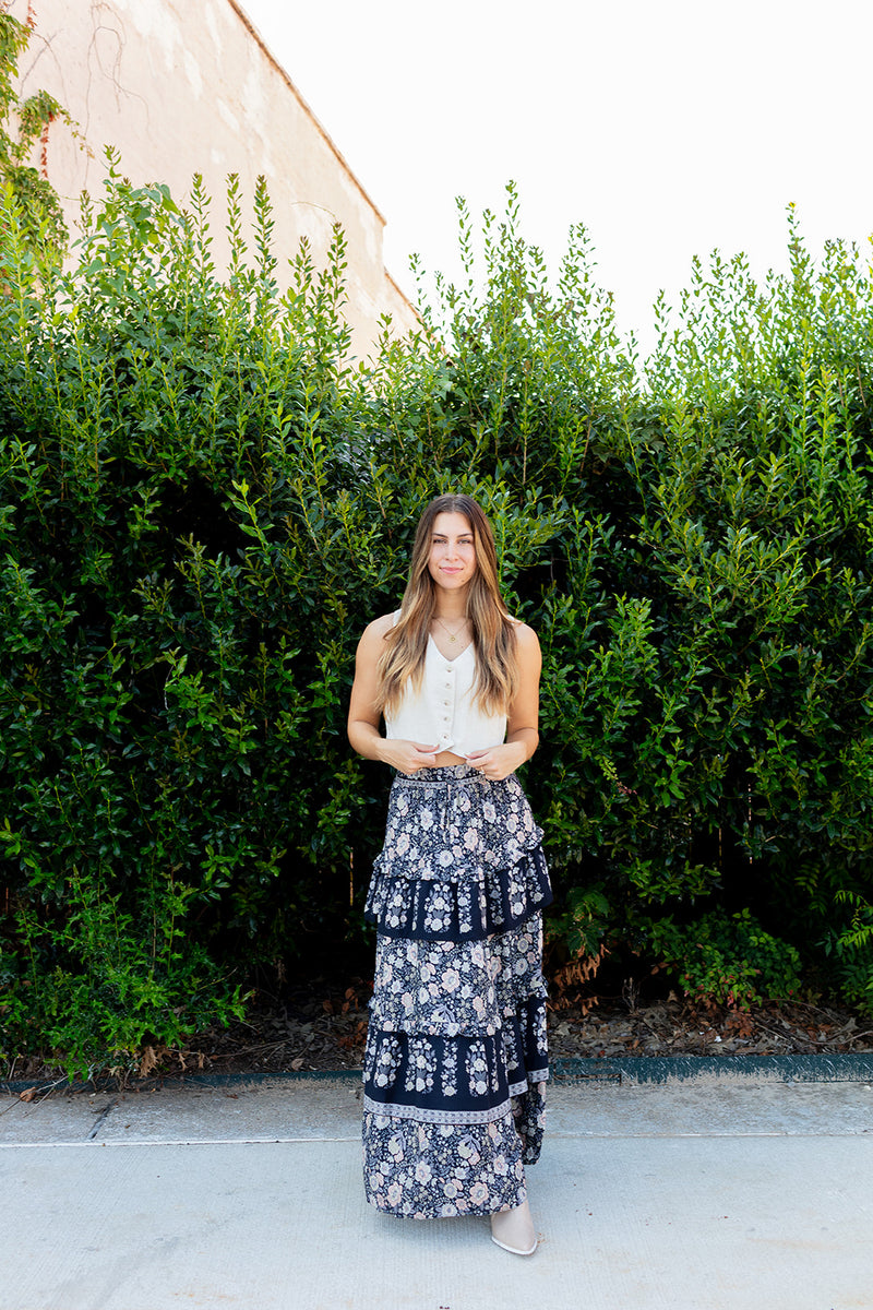 Woman in a white top and floral skirt standing in front of a green hedge.