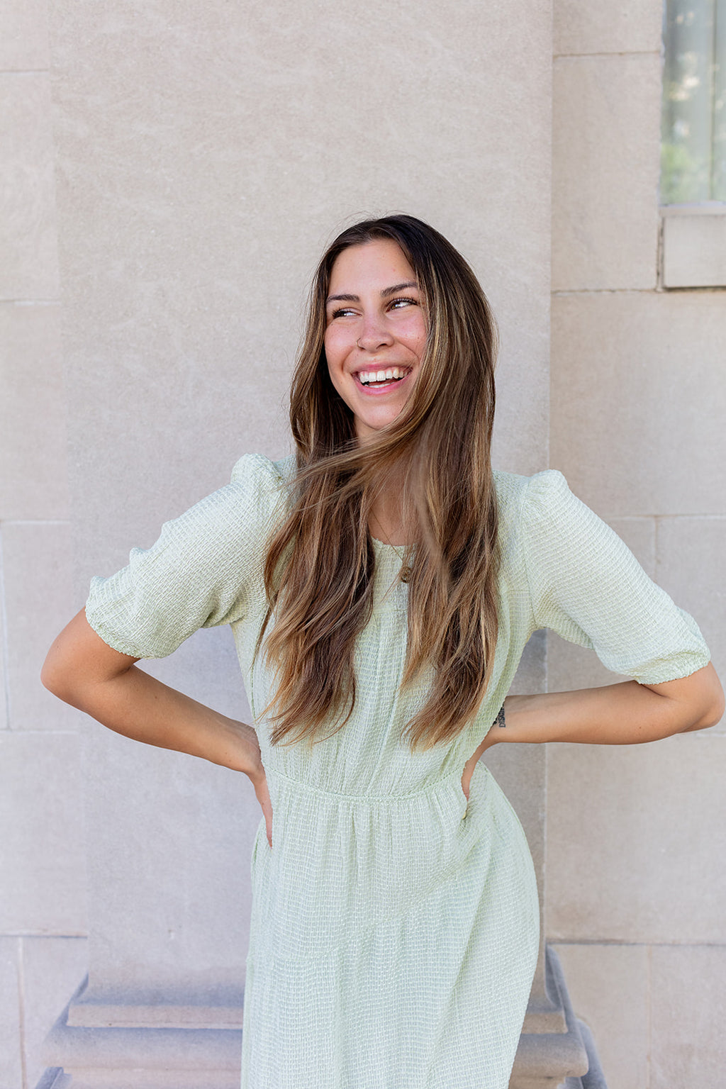 Woman in a light green dress standing against a beige wall.